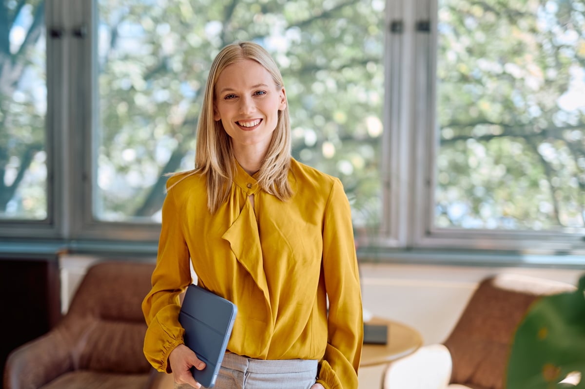 Young professional blonde woman standing in bright office, holding a digital tablet and smiling confidently at the camera