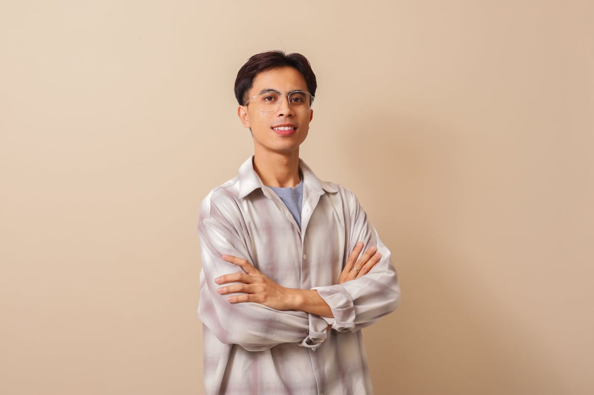 Smiling confident young Asian man standing with crossed arms looking at the camera, isolated on brown background in studio portrait with casual style and friendly expression.